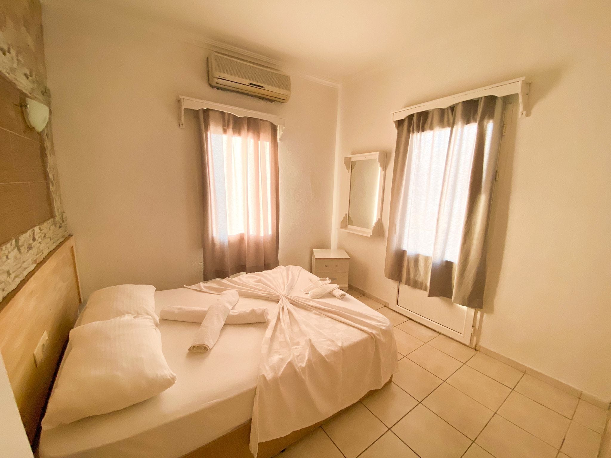 A hotel room with a bed, a bedside table, an air conditioning unit, and two windows with curtains.