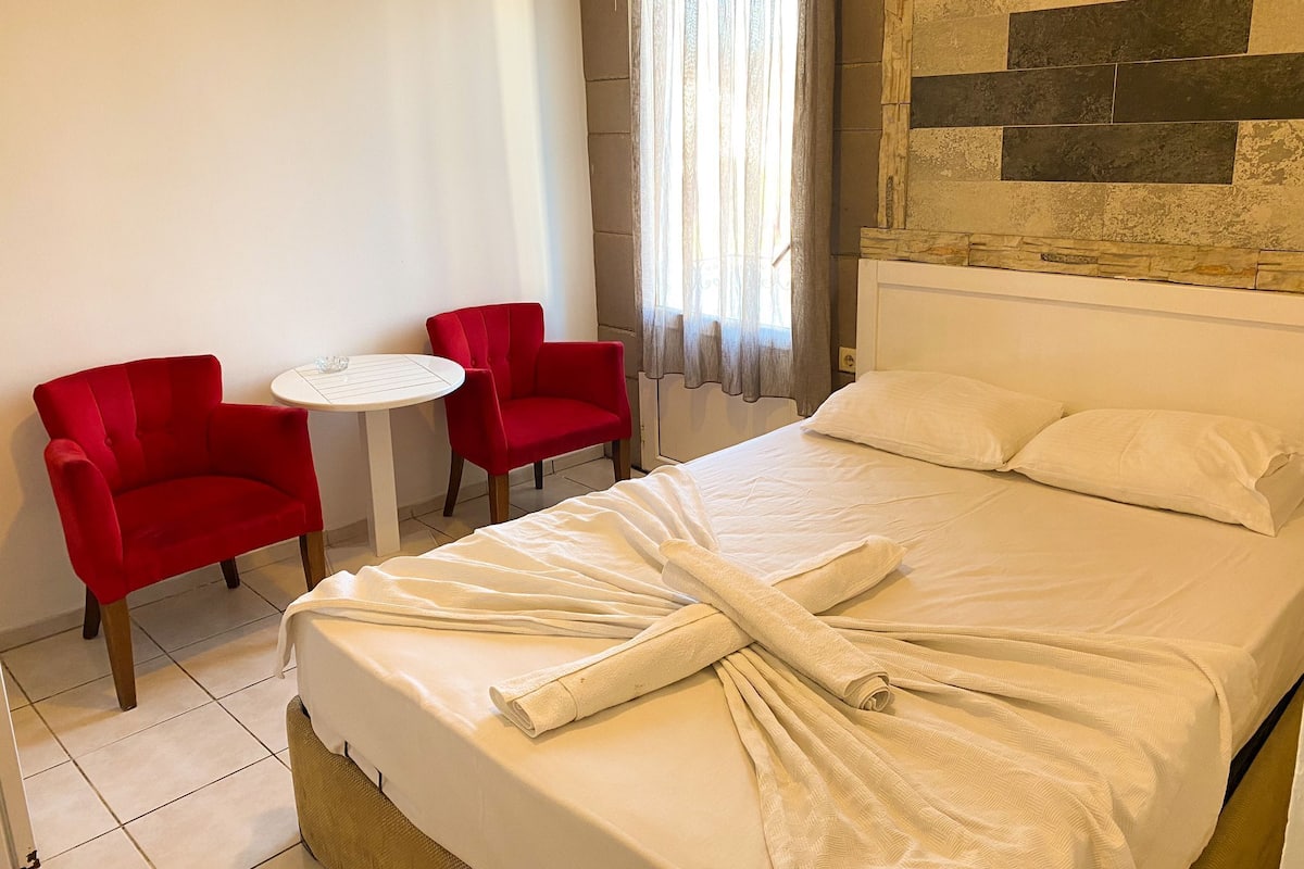 Habitación de hotel con cama, dos sillas rojas, una mesita redonda y una ventana con cortinas.