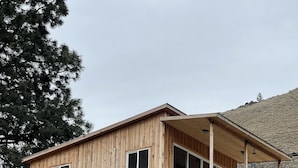 A wooden cabin with a covered porch, large windows, and a view of a hillside. - Hilltop riverview bunkhouse (Riggins)