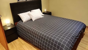 A neatly made bed with a dark checkered bedspread and two white pillows, flanked by two bedside tables with lamps, in a room with a wooden floor and a large abstract painting on the wall.