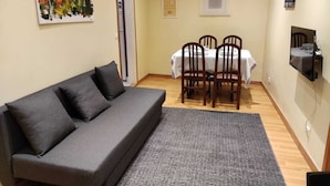 A living room with a grey sofa, a dining table with four chairs, a TV mounted on the wall, and a painting on the wall.