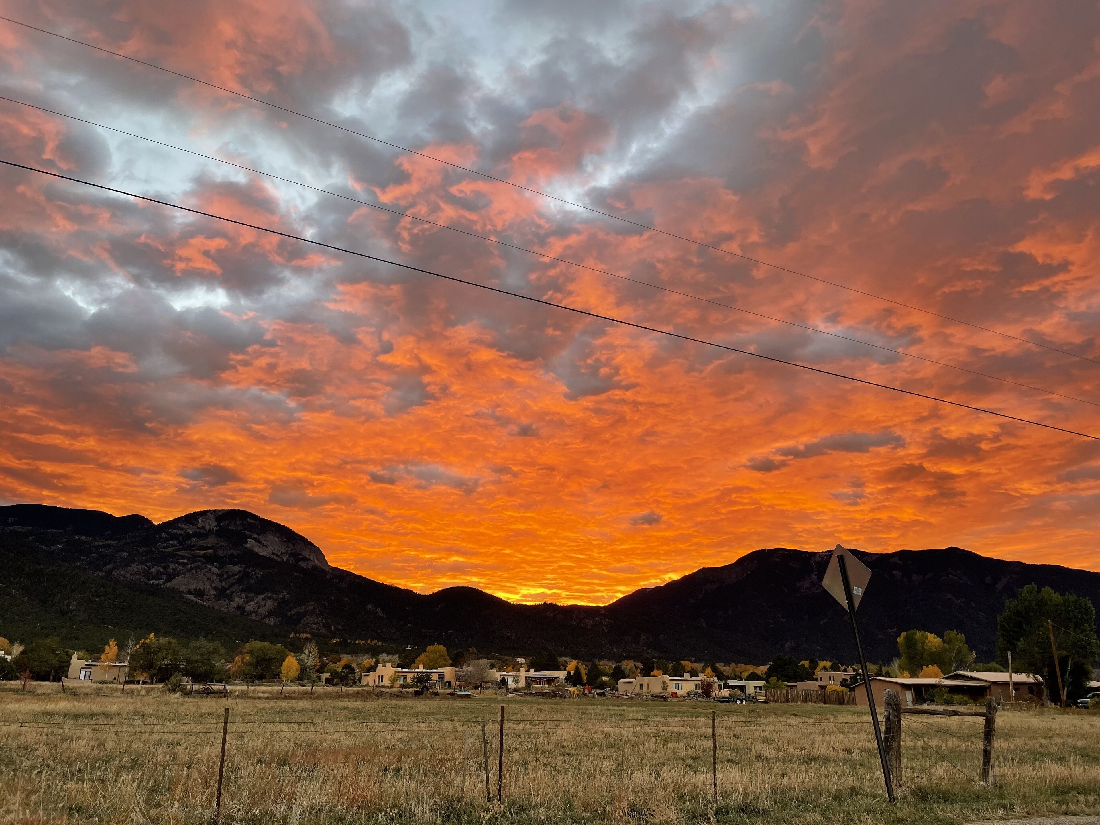 Un paysage rural avec un coucher de soleil, des montagnes en arrière-plan et un petit village composé de maisons et d’une clôture.
