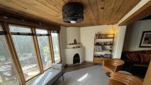 A cozy living room with a fireplace, a bookshelf, and a bench by the large windows.