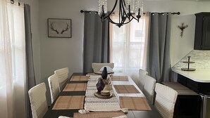 A dining area with a long table, chairs, a chandelier, and a kitchen area visible in the background.