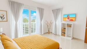 Une chambre moderne avec un grand lit, un téléviseur posé sur un meuble blanc et une fenêtre avec des rideaux.