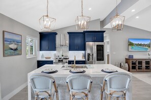 Une cuisine moderne avec des meubles bleu foncé, un réfrigérateur en acier inoxydable et un îlot central doté d’un comptoir blanc. Trois chaises en osier sont disposées autour de l’îlot, et des luminaires suspendus sont accrochés au-dessus.