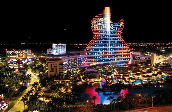 Un hôtel dont la façade extérieure a la forme d’une guitare et est illuminée de lumières rouges et bleues, entouré d’une piscine et de palmiers.