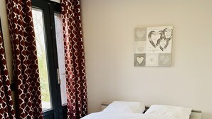 A neatly made bed with a grey and white duvet, a wooden floor, and red patterned curtains. - Pleasant duplex, air conditioning and garden (Montmorency)