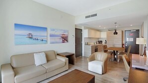 Un salon moderne avec un canapé, une table basse et un coin repas. Deux tableaux ornent le mur, on aperçoit une cuisine au fond et une porte donne accès à l’extérieur.