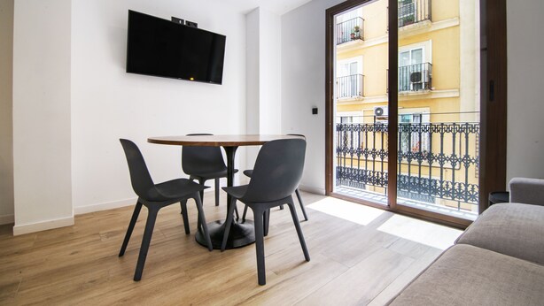 A modern living room with a wooden floor, a round table with four chairs, a flat-screen TV mounted on the wall, and a sliding glass door leading to a balcony with a metal railing. - VLC HOST - Eixarchs 2 (València)