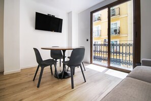 Un soggiorno moderno con pavimento in legno, un tavolo rotondo con quattro sedie, un televisore a schermo piatto montato a parete e una porta scorrevole in vetro che dà su un balcone esterno.