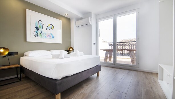 A modern bedroom with a large bed, a bedside table with a lamp, a wall-mounted air conditioner, and a balcony with a railing. - VLC HOST - Eixarchs 5 (València)