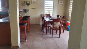 A dining area with a table and chairs, a window with blinds, and a kitchen area with a bicycle and framed pictures on the wall. - Quiet, Spacious and Family Oriented (ANGUILLA)