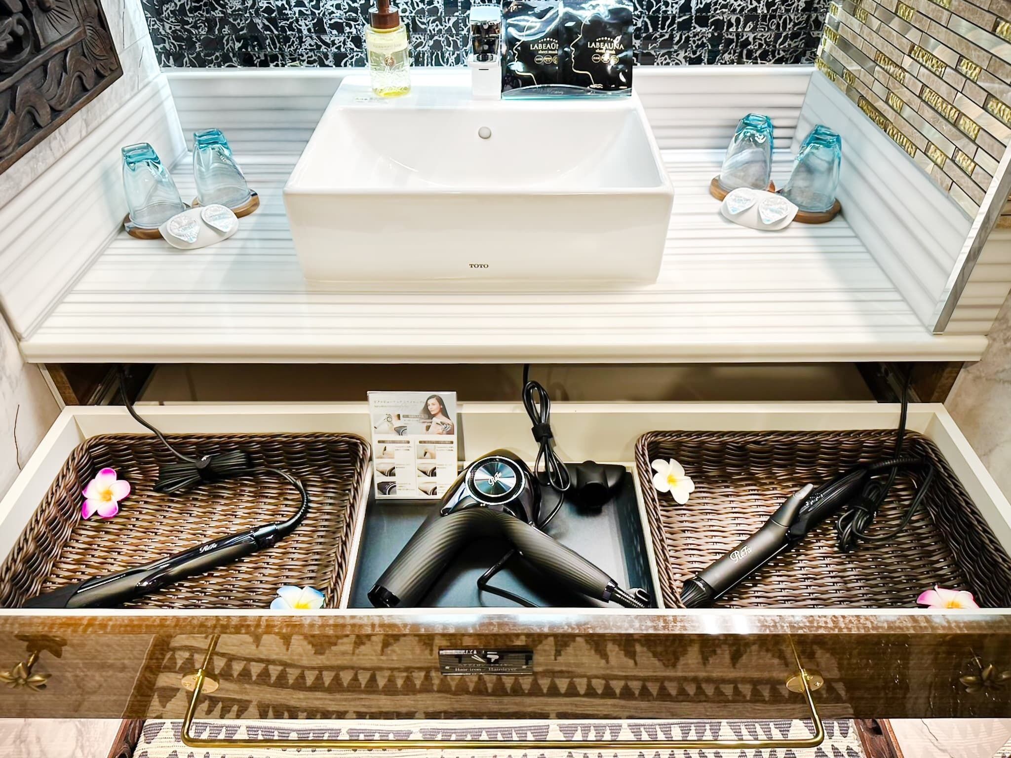 Un baño con un lavamanos blanco, dos botellas de vidrio sobre el mostrador y un espejo encima. Abajo, hay dos cestos de mimbre con utensilios para el cabello y una pequeña caja.