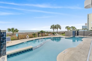 A swimming pool with a view of the ocean, palm trees, and a fenced area with lounge chairs. - The Oasis: Modern 2BR Oceanfront condo with Pool (Myrtle Beach)