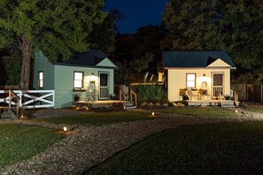 Dos cabañas pequeñas con iluminación exterior, una terraza de madera y un camino de grava.