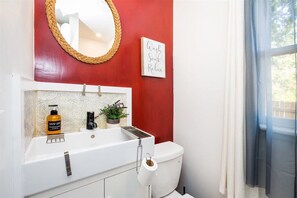 Un baño con lavamanos blanco, pared roja, espejo, inodoro y ventana con cortinas.