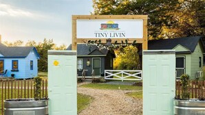 Un senyal de fusta amb la inscripció 'Cape Charles Tiny Livin', un camí de grava que condueix a una porta blanca, i una casa blava al fons.