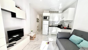 Een modern appartement met een zithoek, inclusief een flatscreen-tv, een bank en een klein tafeltje met een afstandsbediening. De keuken heeft witte kasten, een magnetron en een koelkast.
