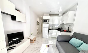 A modern apartment with a living area featuring a flat-screen TV, a sofa, and a small table with a remote control. The kitchen has white cabinets, a microwave, and a refrigerator.