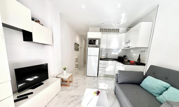 A modern apartment with a living area featuring a flat-screen TV, a sofa, and a small table with a remote control. The kitchen has white cabinets, a microwave, and a refrigerator.