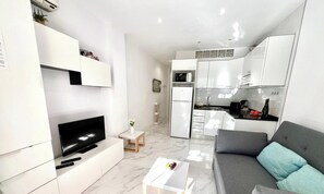 A modern living room with a flat-screen TV, a sofa, a coffee table, and a kitchen area with white cabinets and appliances.