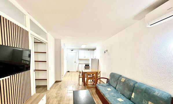 A living room with a TV, a sofa, a dining table with chairs, and a kitchen in the background.