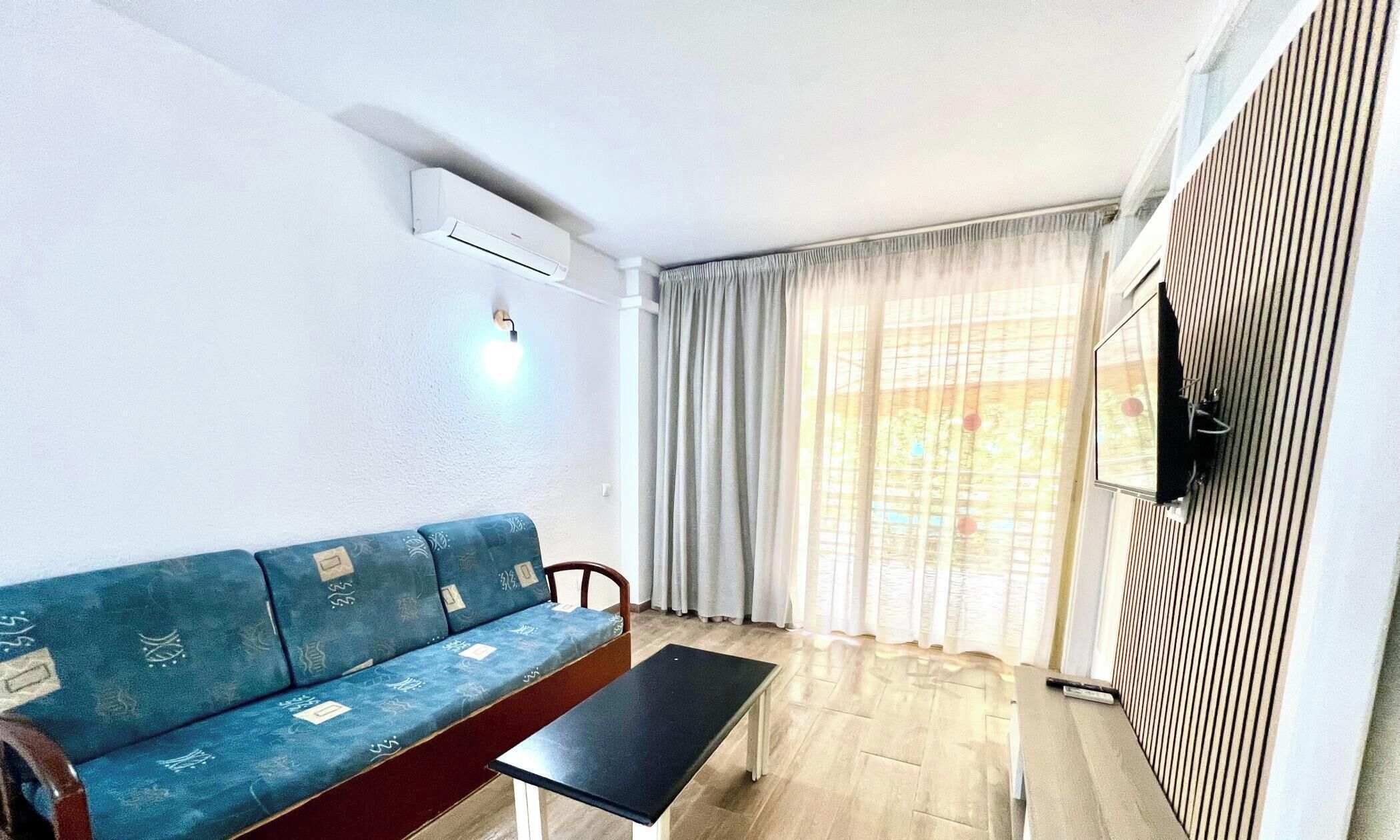 A room with a sofa, a table, a TV mounted on the wall, and a window with curtains.