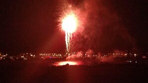 Una escena nocturna con un brillante espectáculo de fuegos artificiales sobre una masa de agua, con luces de la ciudad a lo lejos.