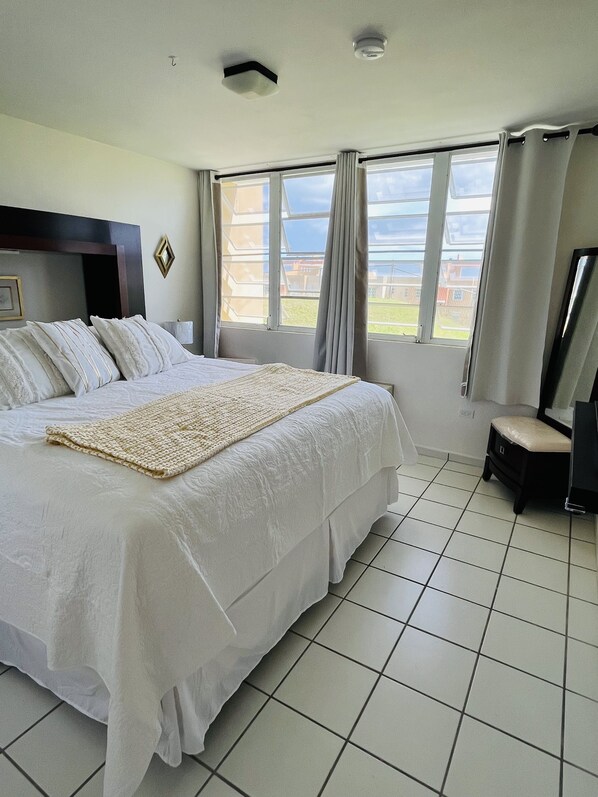 Una cama grande con sábanas blancas y un plaid tejido, un taburete pequeño y una ventana con cortinas.