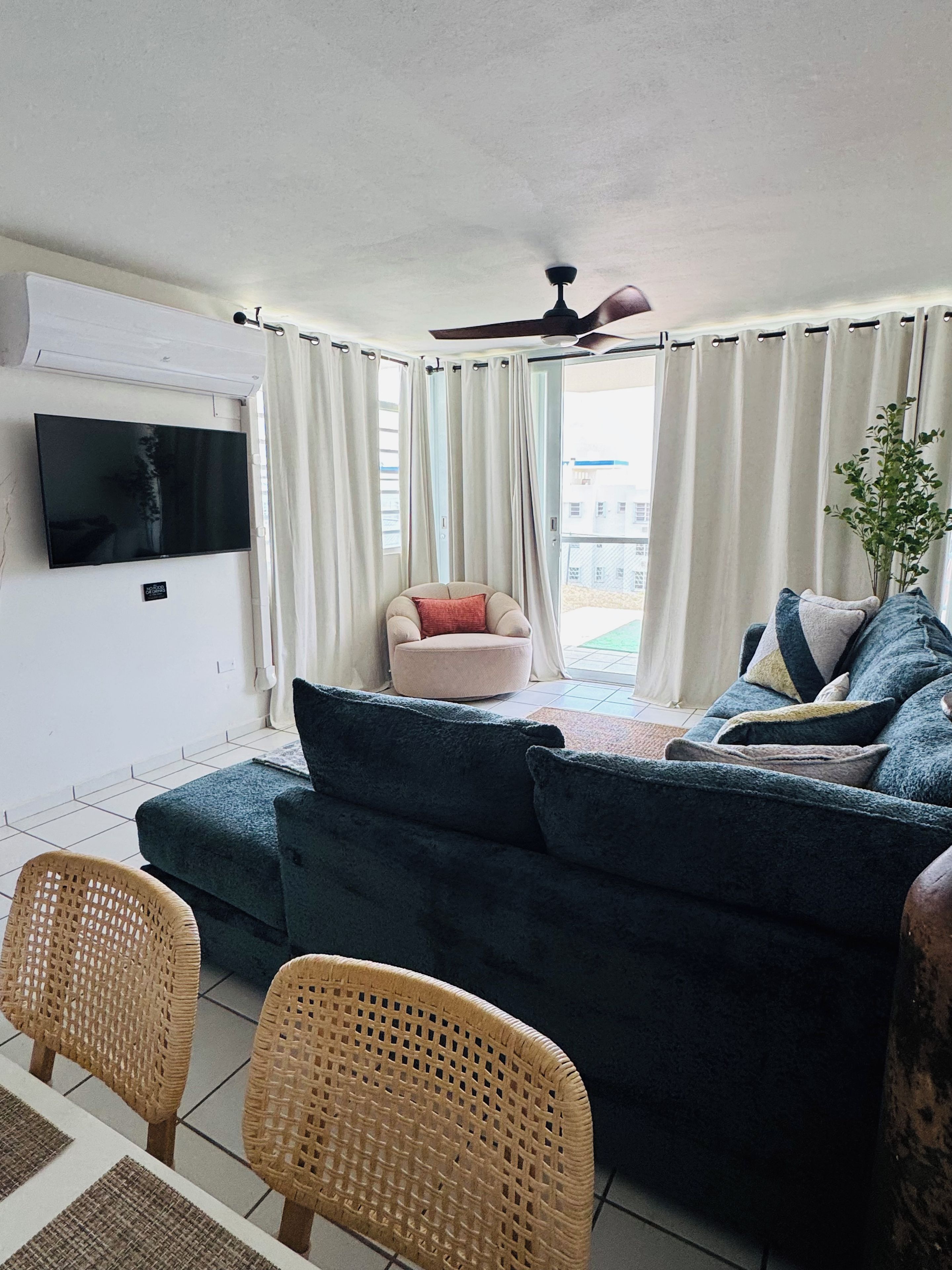Un salon avec un canapé bleu foncé, un téléviseur fixé au mur, un ventilateur de plafond et un coin repas avec des chaises en osier.