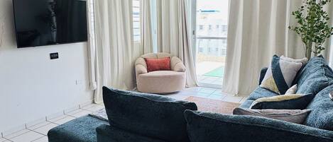 Un salon avec un canapé bleu foncé, un téléviseur fixé au mur, un ventilateur de plafond et un coin repas avec des chaises en osier.