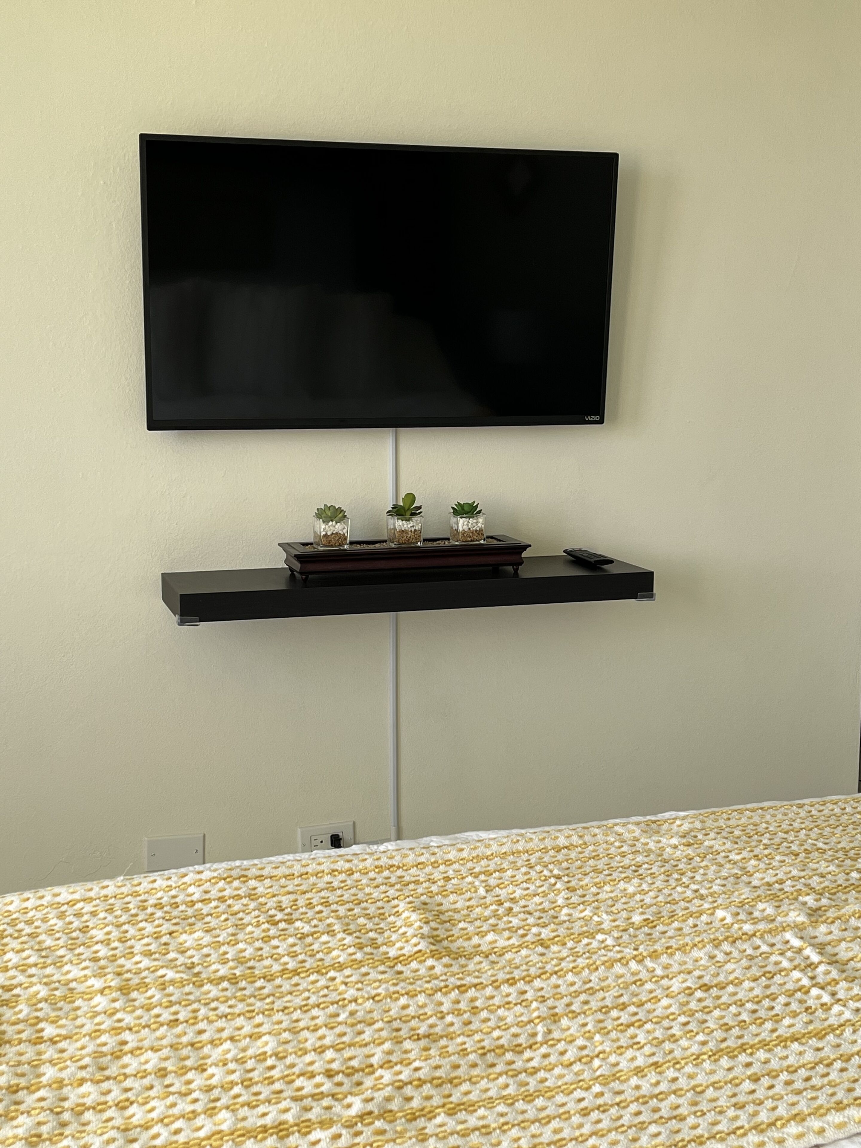 Une pièce avec un téléviseur à écran plat fixé au mur, une petite étagère en dessous où sont posées trois plantes en pot, et un lit recouvert d’une couvre-lit à motifs.
