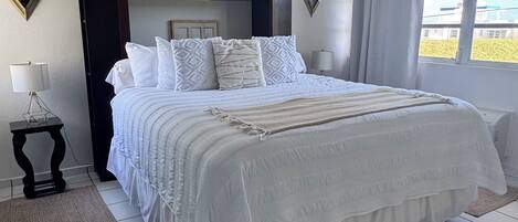A neatly made bed with white bedding, a dark headboard, and a view of the outdoors through a window.