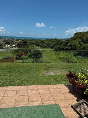 A view of a grassy area with a fence, a paved walkway, and a distant view of buildings and hills. - Spacious 3 bedroom apartment with sea view short drive to ferry station. (Ceiba)