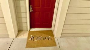 Eine rote Tür mit weißem Rahmen, eine Fußmatte mit der Aufschrift "Welcome" und eine Wandleuchte.