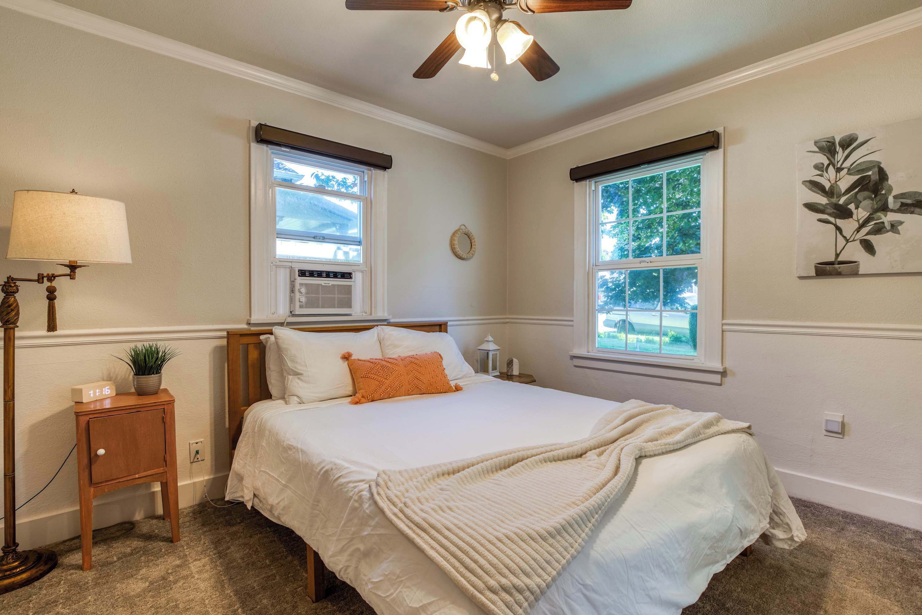 A bedroom with a bed, bedside tables, a nightstand, a lamp, a ceiling fan, and two windows with curtains.