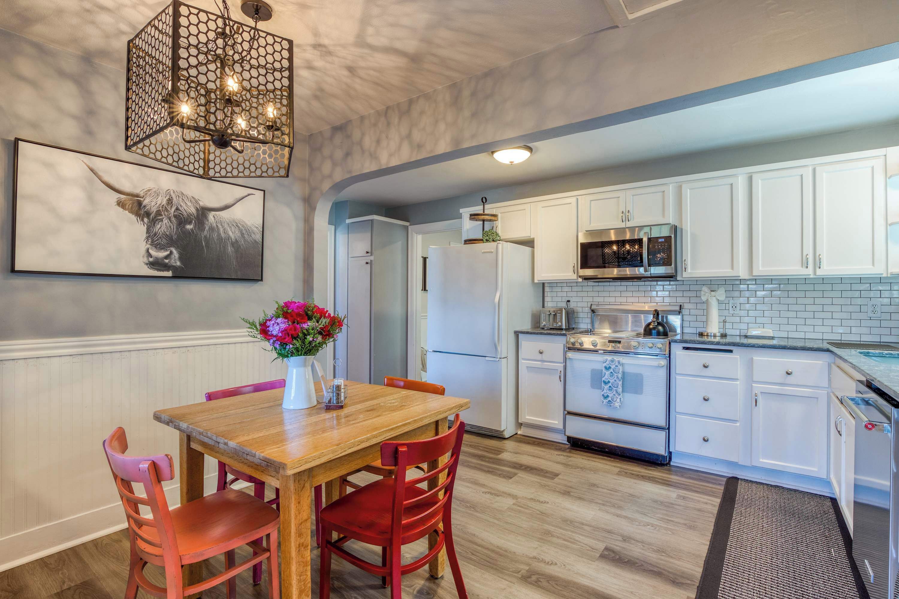 A kitchen with white cabinets, a wooden dining table with red chairs, a chandelier, and a bull-themed artwork.