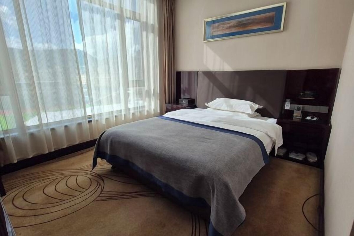 Une chambre d’hôtel avec un lit, une table de chevet, une lampe et une fenêtre avec des rideaux légers.