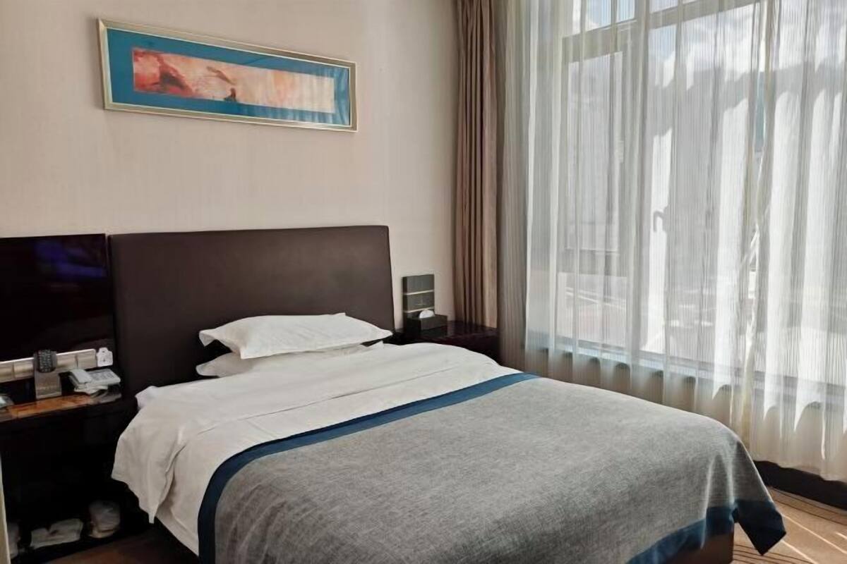 A hotel room with a large bed, a bedside table with a phone and a lamp, a painting on the wall, and a window with sheer curtains.