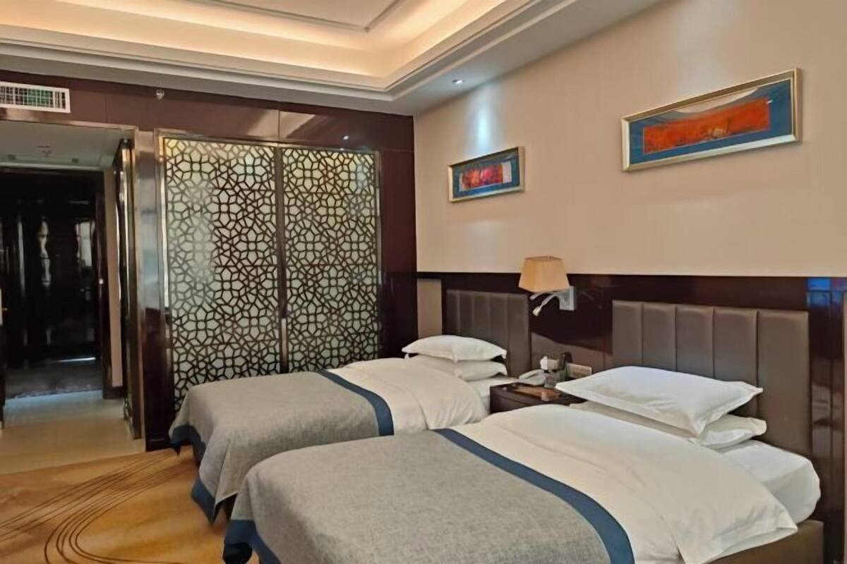 A hotel room with two beds, a decorative partition, a painting, and a nightstand with a lamp.