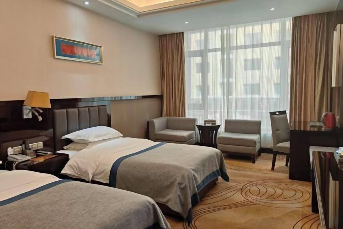 A hotel room with two beds, a desk, a chair, a lamp, and a large window with curtains.