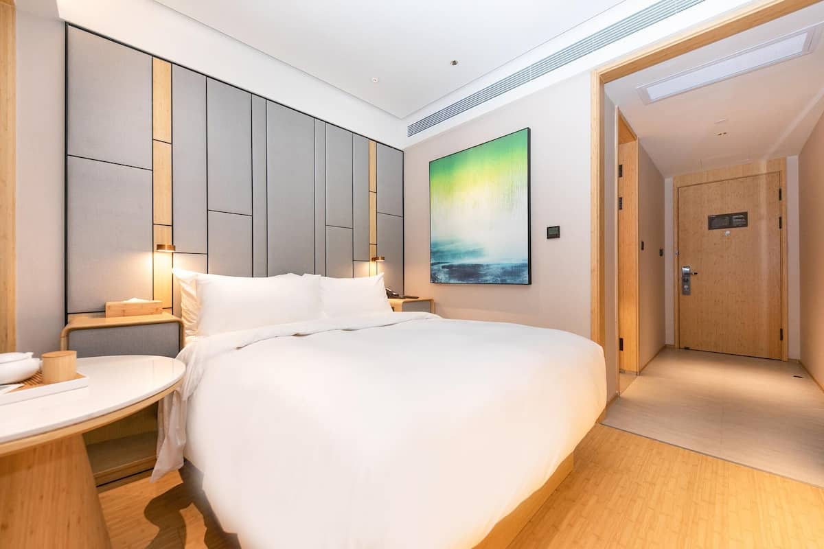 A modern hotel room with a large bed, a bedside table, a nightstand, a wall-mounted TV, and a door to the bathroom.
