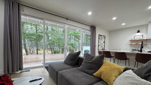 Una sala de estar moderna con un sofá gris, almohadones blancos y amarillos con estampado, un control remoto sobre una mesa blanca y vistas a un bosque a través de puertas de cristal correderas grandes.