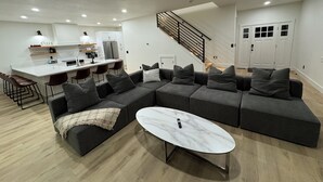 Una sala moderna con un sofá modular, una mesa de centro de mármol y una cocina con armarios blancos y un taburete de bar.