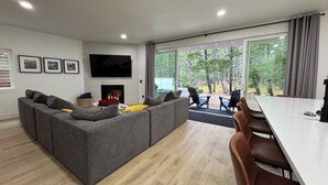 Una sala de estar moderna con un sofá seccional gris, una chimenea, un televisor de pantalla plana y una zona de comedor con una mesa blanca y sillas marrones.