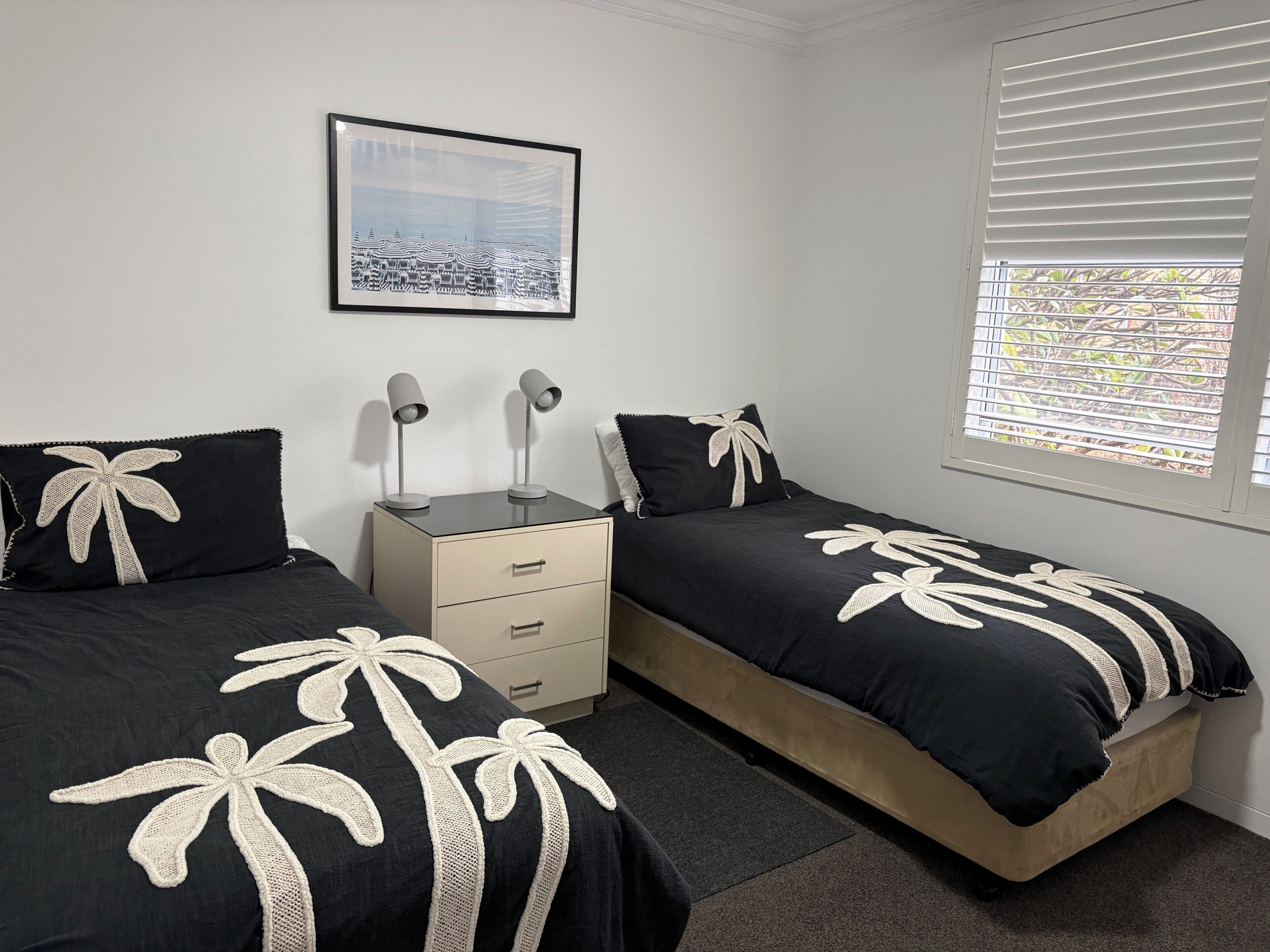 A bedroom with two beds, a nightstand, a drawer unit, a framed picture on the wall, and a window with blinds.