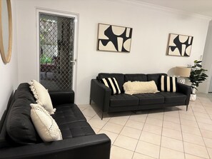 A living room with a black sofa, a black armchair, and a black leather couch. There are abstract paintings on the wall and a potted plant.