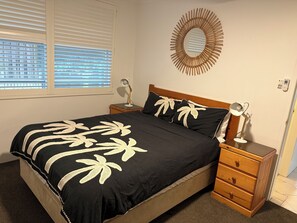 Une chambre à coucher avec un lit, des tables de chevet, un miroir et une fenêtre avec des stores.