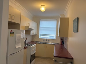 A compact kitchen with white cabinets, a gas stove, a refrigerator, and a window with blinds.
